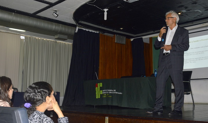 Gualter Nunes Maia fala ao microfone, de pé no palco do auditório, com apresentação de slides ao fundo