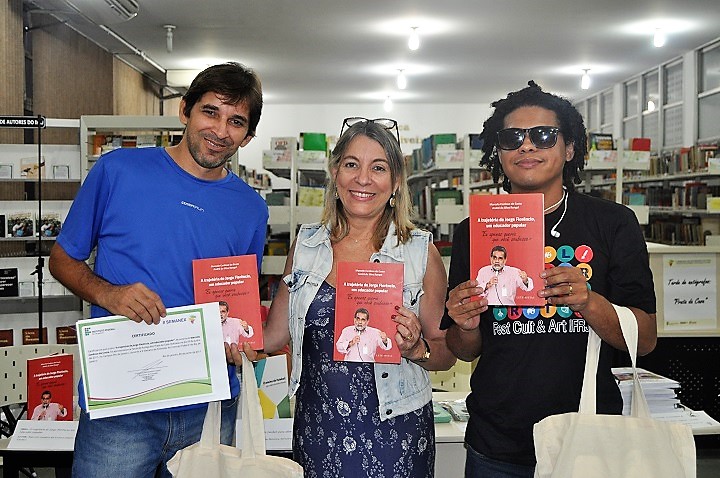 Três pessoas posam, na biblioteca, com exemplares do livro do professor Marcelo nas mãos 