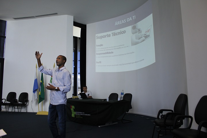 Palestrante, de pé, fala sobre área de TI com apresentação de slides atrás