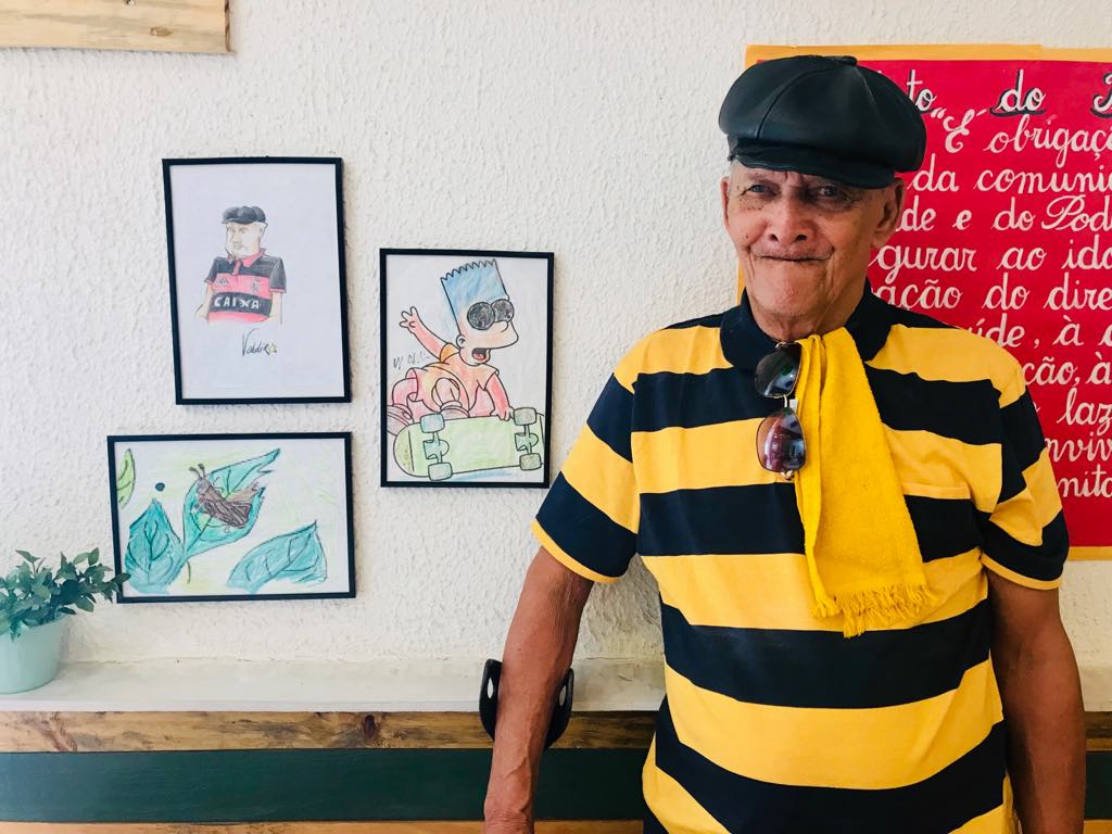 Homem idoso posa para foto ao lado de quadros desenhados a lápis na parede