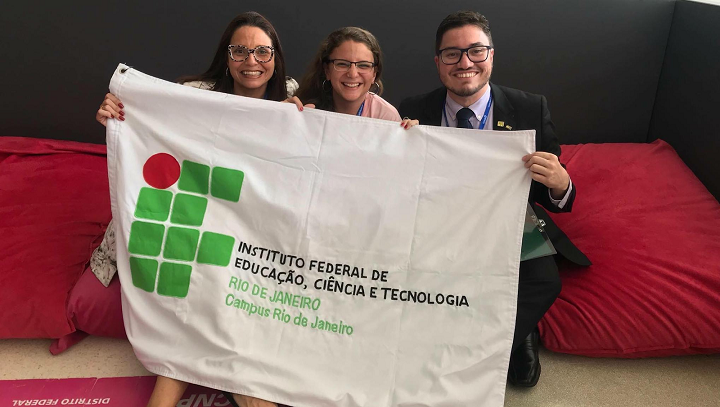 Alessandra, Maira e Rafael sentados em um sofá vermelho e segurando a bandeira do IFRJ