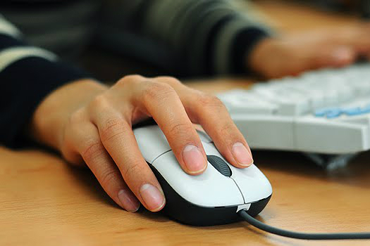 close de uma mão mexendo num mouse com teclado de computador ao fundo