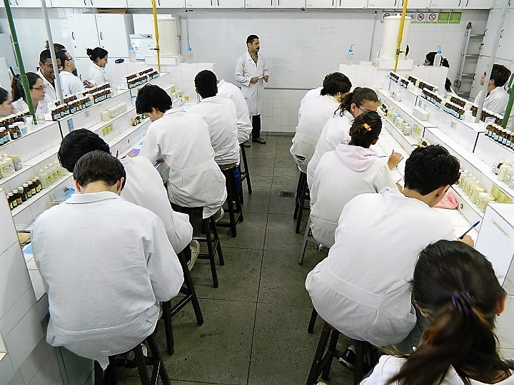 Alunos tendo aula em laboratório, cercados por vidros de itens químicos, com professor à frente da sala