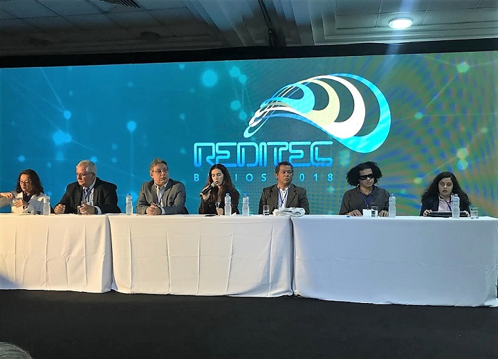Integrantes da mesa-redonda sentados atrás da mesa na frente do auditório, com telão atrás com a logo da Reditec 2018