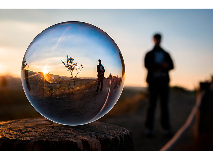 Vemos uma bola de vidro que reflete a imagem de um homem em um campo aberto, com um pequeno arbusto ao lado sob o pôr do sol
