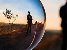 Vemos uma bola de vidro que reflete a imagem de um homem em um campo aberto, com um pequeno arbusto ao lado sob o pôr do sol