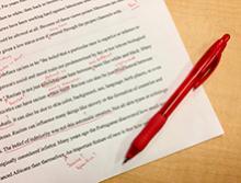 Vemos um texto impresso escrito na língua inglesa com correções em vermelho feitas pela caneta que se encontra deitada na lateral direita do papel